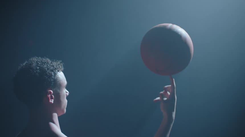 A cinematic shot of a young, shirtless basketball player spinning a ball on his finger in a dark, hazy room