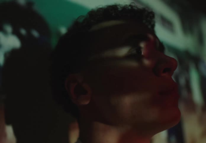 A contemplative young man's face is lit up by a movie projector in a dark, cinematic shot