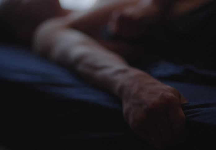 A man's hand comes into focus as he lies shirtless in a dimly lit bed