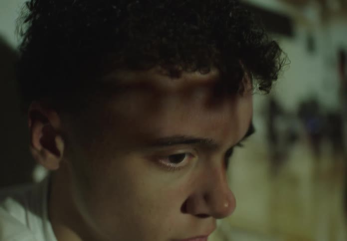 A serious young man watches a film projection of a basketball game, with the images flickering across his face in a dark room