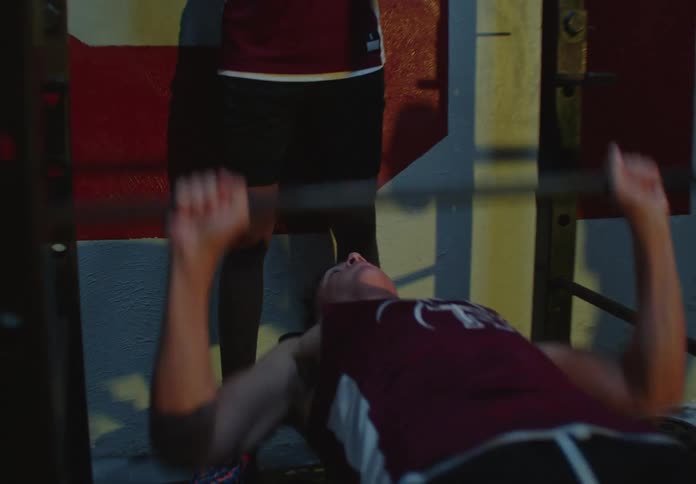 Two basketball players strength train together, with one spotting the other during a bench press in a dimly lit gym
