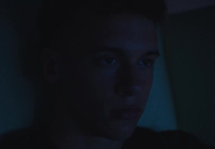 A young man intently watches a screen, his face illuminated by its blue glow in a dark room