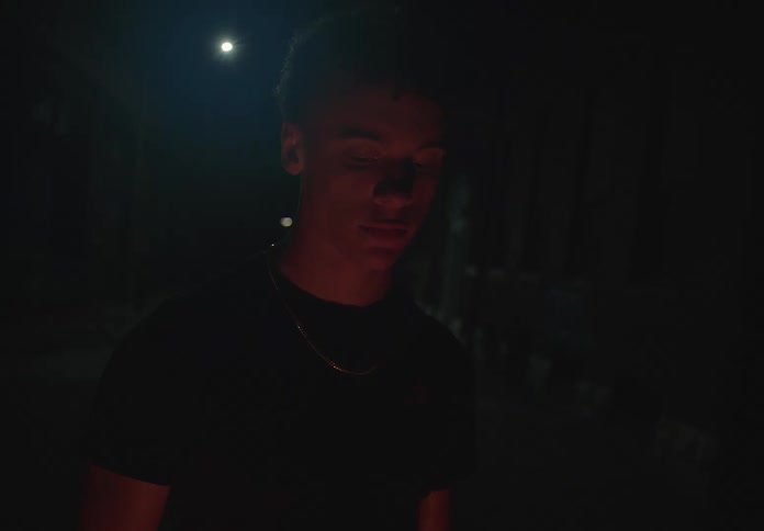 A contemplative young man stands alone in a dark urban alley at night, his face illuminated by a warm, unseen light from below