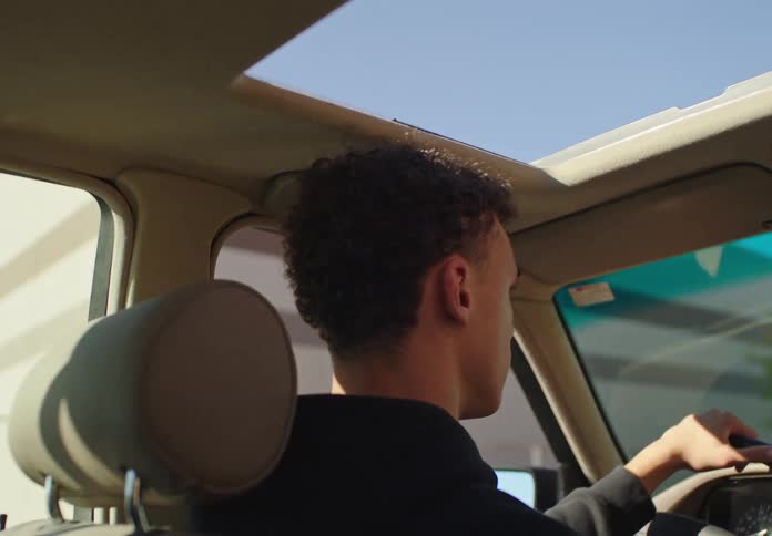 Over-the-shoulder shot from the backseat shows a young man with curly hair driving on a sunny day