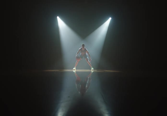 Wide shot of a basketball player dribbling on a dark court under dramatic spotlights