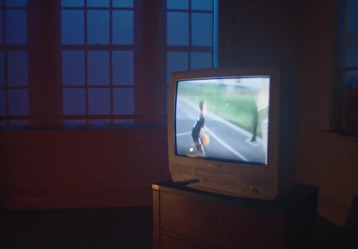 An old CRT television plays nostalgic home videos of a childhood basketball game and practice in a dimly lit, blue-hued room