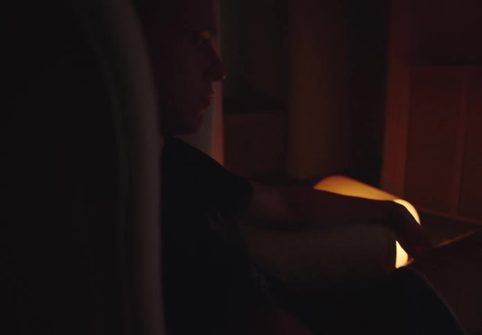 A young man sits in a dark, atmospherically lit room watching a sports game on an old television