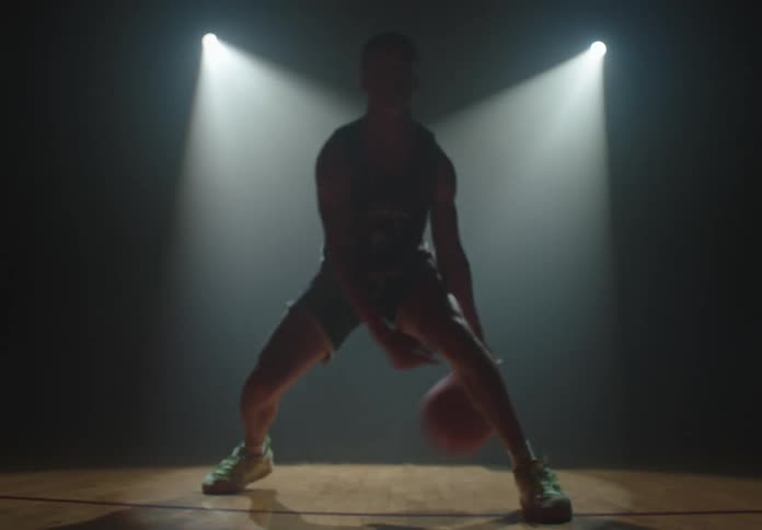 A basketball player practices dribbling between his legs in a dark, hazy gym, dramatically silhouetted by bright spotlights