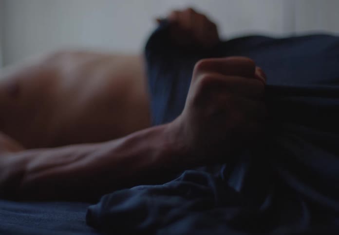 A shirtless man's hand clenches the bedsheets in slow motion in a dimly lit bedroom
