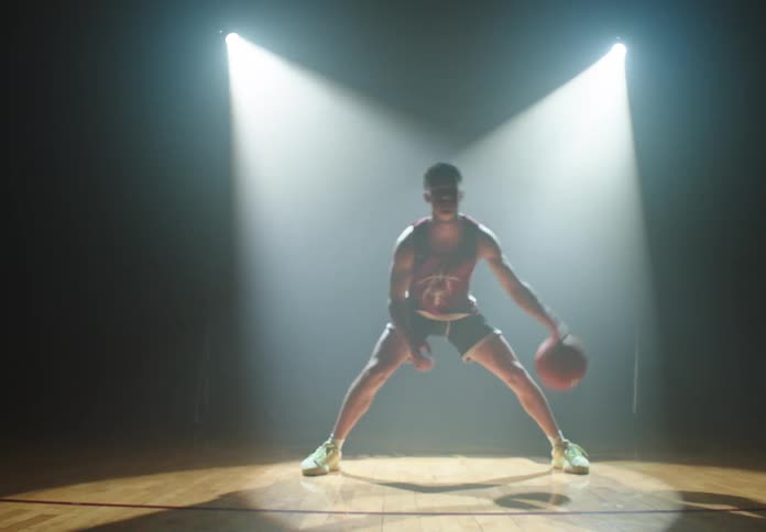 A focused basketball player intensely dribbles on a dark, reflective court under dramatic spotlights as the camera slowly pushes in