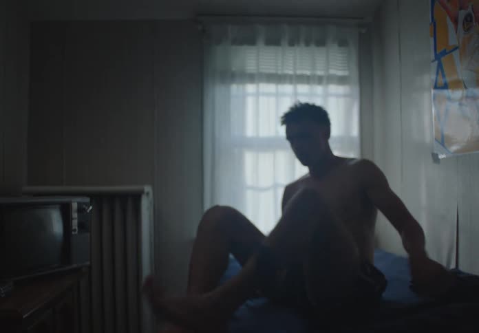 A pensive young man slowly sits up on the edge of his bed in a dimly lit, moody bedroom