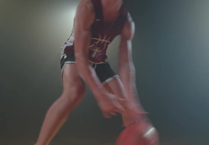 A determined basketball player skillfully dribbles a ball across a court under dramatic spotlights in a dark, hazy gym