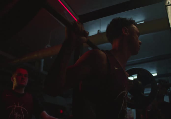 A determined basketball player performs heavy barbell squats in a dark gym while his teammates watch and support him