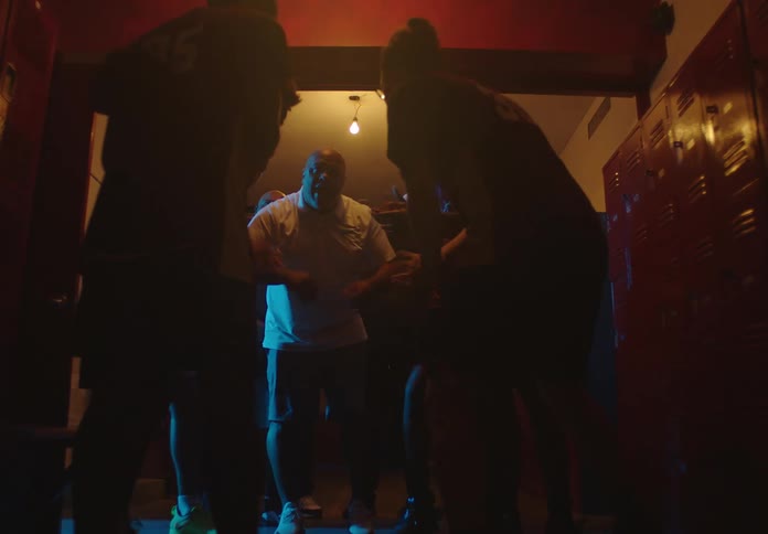 In a dramatic, dimly lit locker room, a coach passionately gives a motivational speech to his basketball team before they huddle together