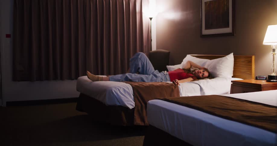 A smiling young woman relaxes on a bed in a dimly lit motel room at night