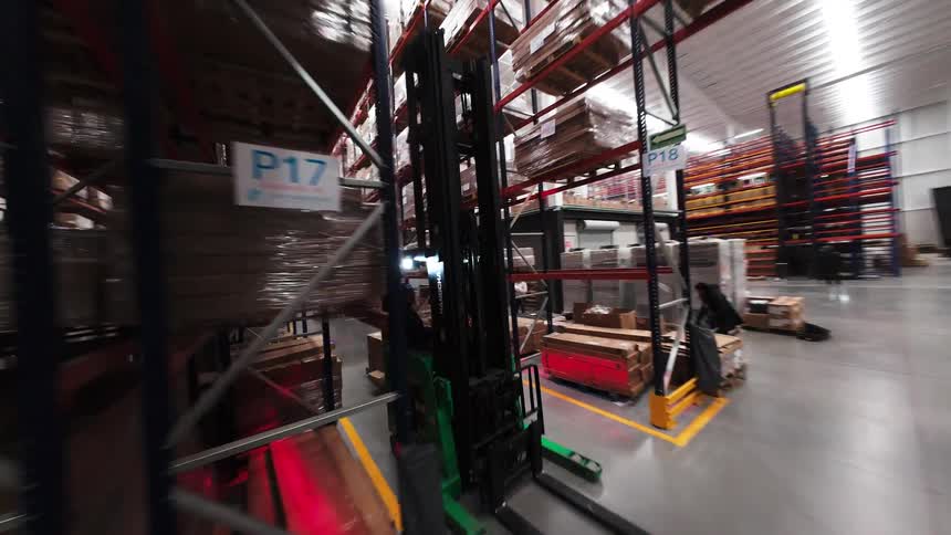 Dynamic drone footage moves backward through a vast industrial warehouse aisle, showcasing organized shelves stacked high with boxed inventory and workers operating a forklift