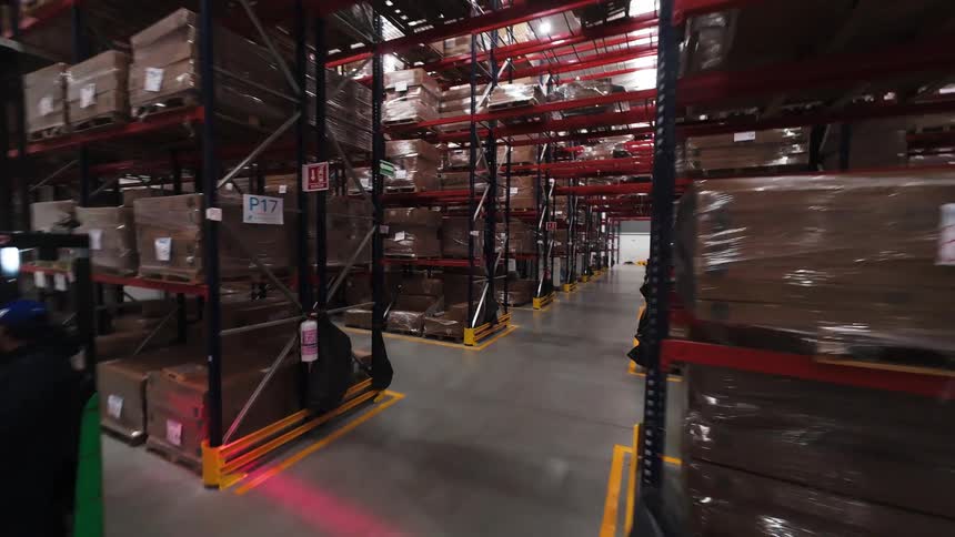 A tracking shot moves through a bustling warehouse as workers operate forklifts and pallet jacks among towering shelves of inventory