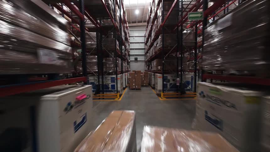 A fast-moving point-of-view shot travels down a long aisle in a logistics warehouse stacked with pallets and boxes
