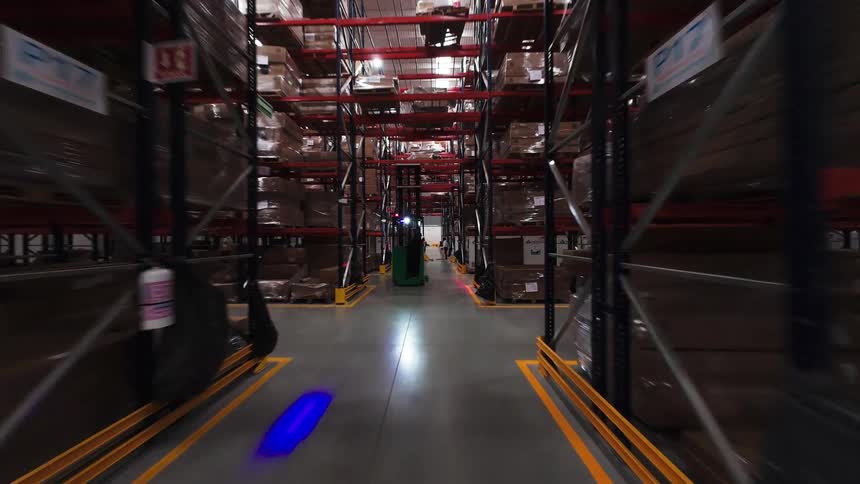 FPV drone footage flies through a busy warehouse aisle, past tall shelves of inventory and workers