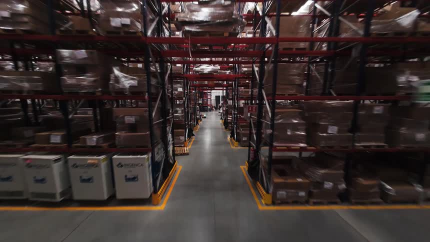 FPV drone flies rapidly down a long aisle between towering shelves packed with goods in a massive industrial warehouse