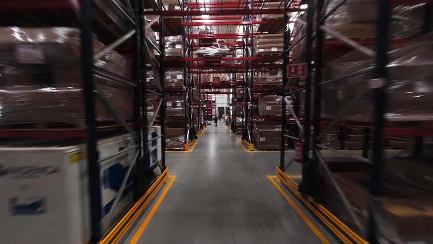 A smooth FPV shot glides down a long aisle in a massive warehouse, passing by tall shelves stacked high with inventory