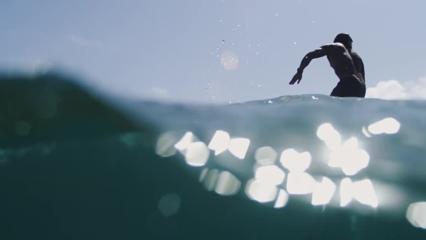 Super slow motion, low-angle shot of a silhouetted surfer on his board as the camera submerges into the dark ocean