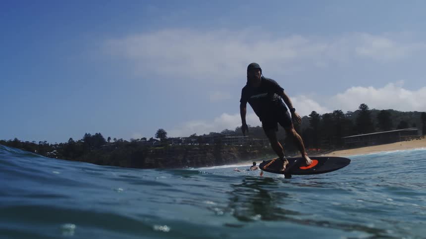 A man on a hydrofoil surfboard smoothly glides across the ocean's surface in stunning slow motion, with the sandy beach in the background