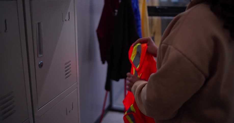 A female worker with curly hair smiles as she takes her orange safety vest from her locker and chats with a colleague before starting her workday