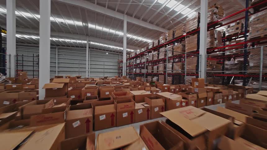 An FPV drone flies through a massive, busy warehouse filled with cardboard boxes, high shelves of inventory, and workers sorting packages