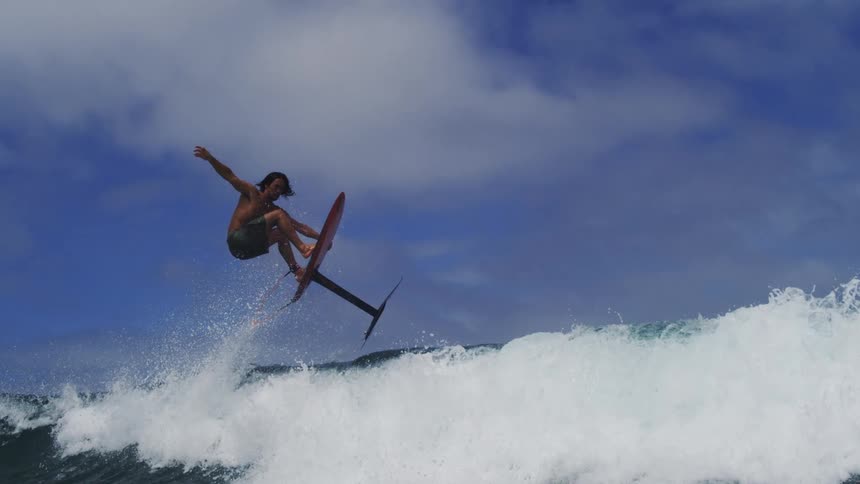 A man on a hydrofoil surfboard expertly launches off a wave and soars through the air in dramatic slow motion