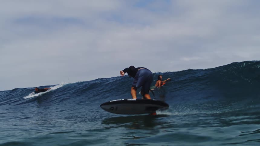 A man skillfully rides a large ocean wave on an electric hydrofoil surfboard in a dramatic slow-motion shot