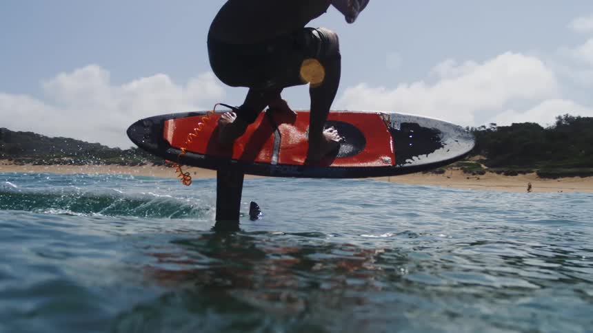 A man with long hair skillfully rides an electric hydrofoil surfboard in stunning slow motion, gliding effortlessly over calm ocean waves towards the beach