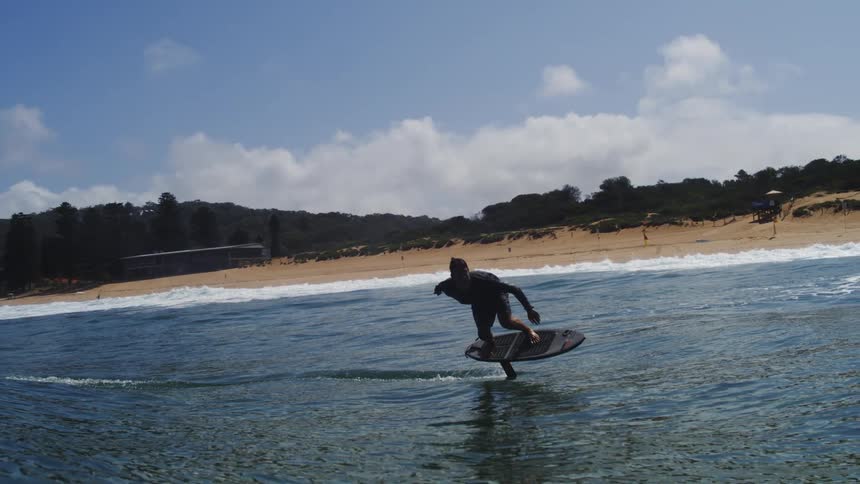Slow motion shot of a man gracefully hydrofoil surfing on the ocean towards a sunny beach