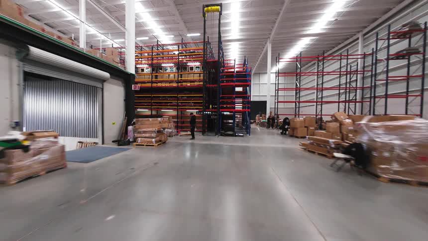 A ground-level first-person view drone navigates through the aisles of a large, busy warehouse, passing by a forklift and flying under shelving