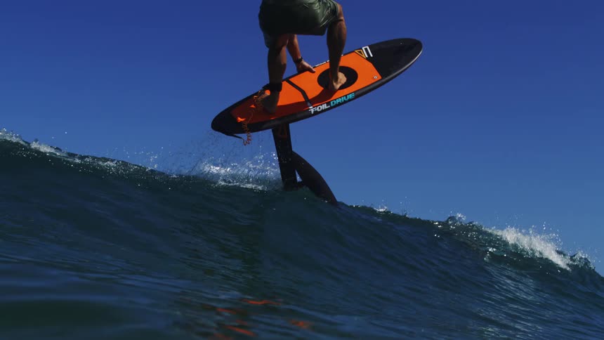 A surfer skillfully rides a hydrofoil board on a wave, eventually launching into the air in a breathtaking slow-motion jump against a clear blue sky