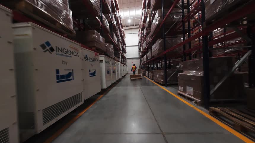 A smooth tracking shot moves through the long aisles of a vast logistics warehouse, with high shelves stacked with boxes and pallets