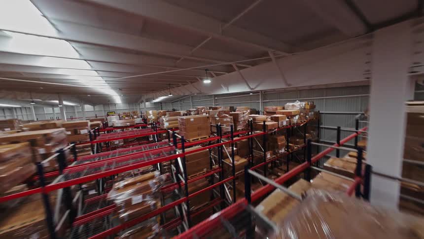An FPV drone shot soars through the aisles of a vast warehouse, revealing endless shelves stacked high with boxes and pallets