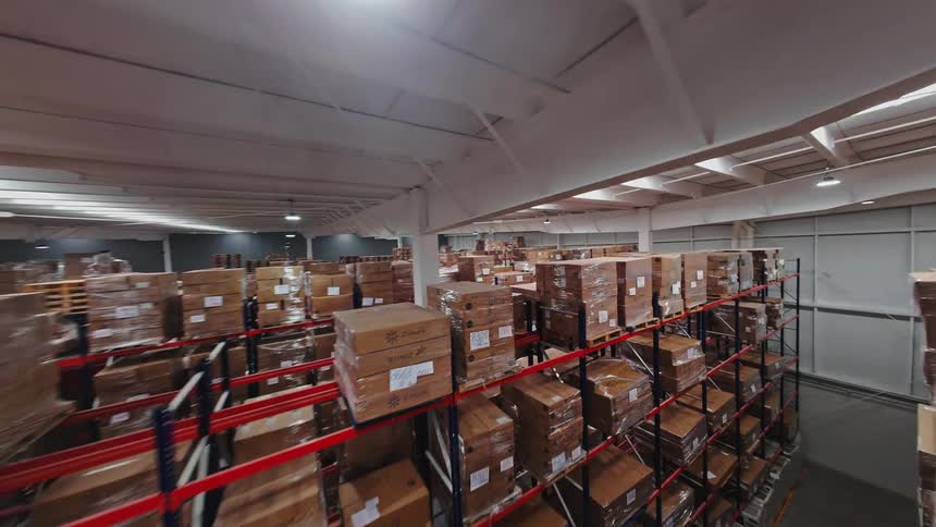 A dynamic FPV drone flies through the aisles and over the tall racks of a vast, brightly lit warehouse filled with inventory