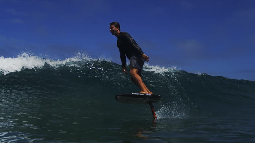 An athletic man expertly surfs a hydrofoil board on a beautiful blue ocean wave in super slow motion