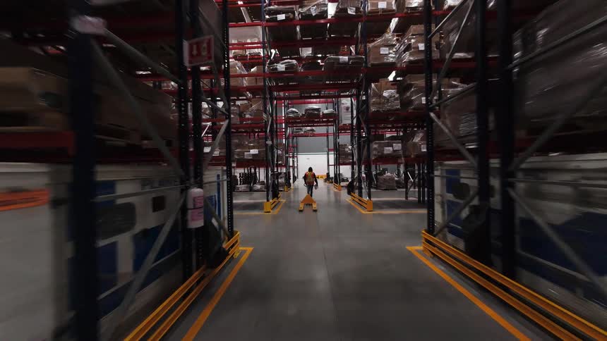 A smooth, forward-moving shot explores the long aisles of a large distribution warehouse with workers operating pallet jacks among towering shelves of merchandise