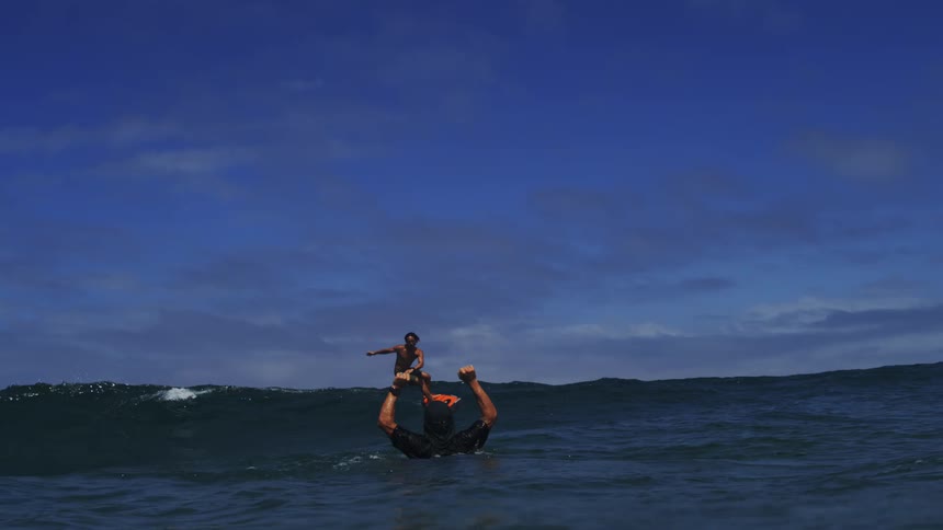 A surfer on a hydrofoil board successfully rides a wave as his friend cheers him on from the water in super slow motion