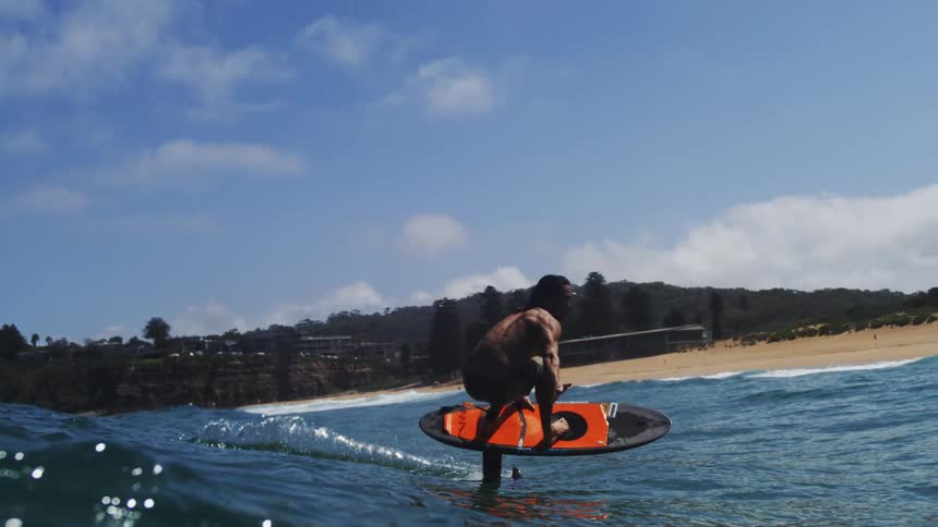 In slow motion, a skilled surfer with long hair glides effortlessly across a blue wave on a hydrofoil board