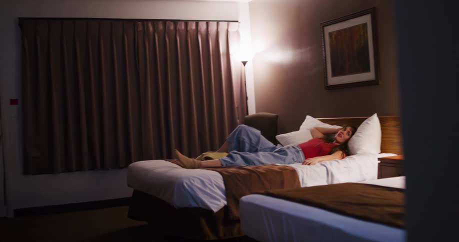 A young woman in casual clothes lies on a bed, resting in a dimly lit hotel room at night