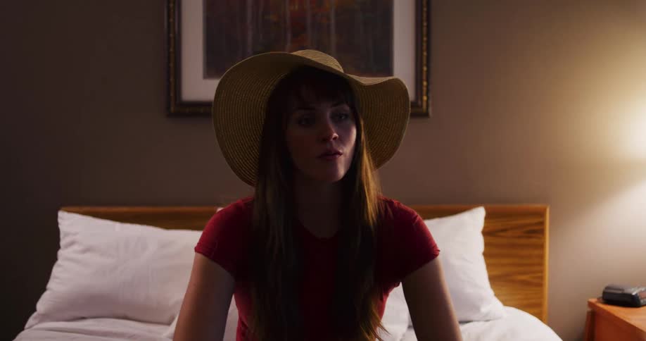 A young woman wearing a red shirt and a large sun hat sits on a hotel bed, her mood shifting from thoughtful to playful