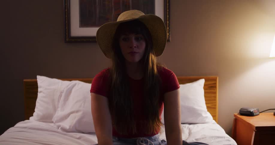 A young woman wearing a sun hat falls backward onto a hotel bed in slow motion and sits up looking contemplative