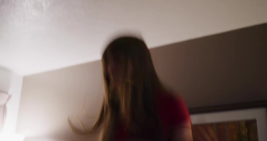 A joyful woman wearing a hat dances and jumps on a bed in a dimly lit room in slow motion