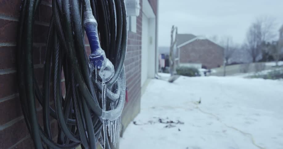 A close-up of a garden hose covered in ice and icicles hanging on a brick wall as snow falls in the background