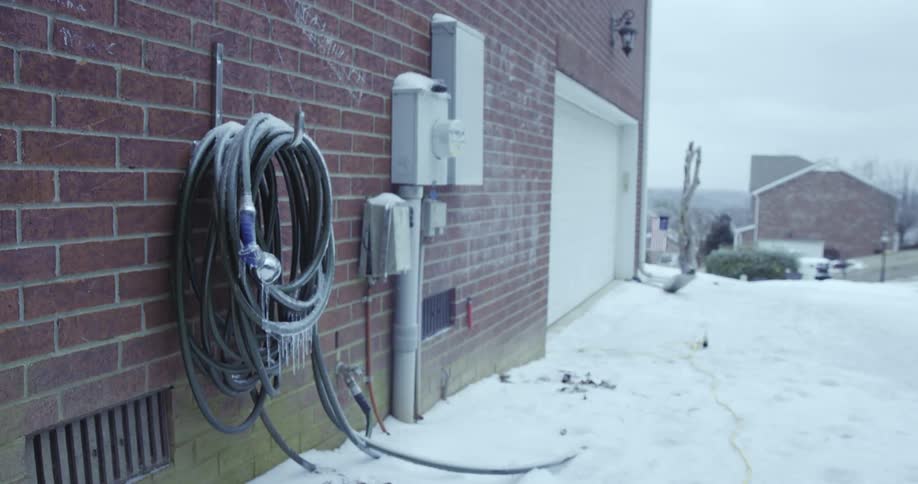 A frozen garden hose with icicles hangs coiled on the brick wall of a house on a snowy winter day