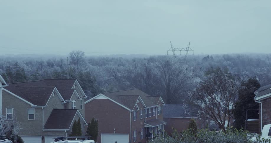 Snow gently falls on a quiet suburban neighborhood with a snow-covered forest and a power line tower in the distance