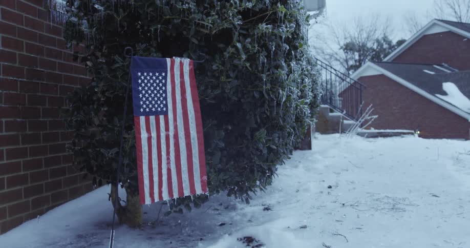 An American flag hangs still in a yard covered in snow and ice as light sleet falls during a winter storm